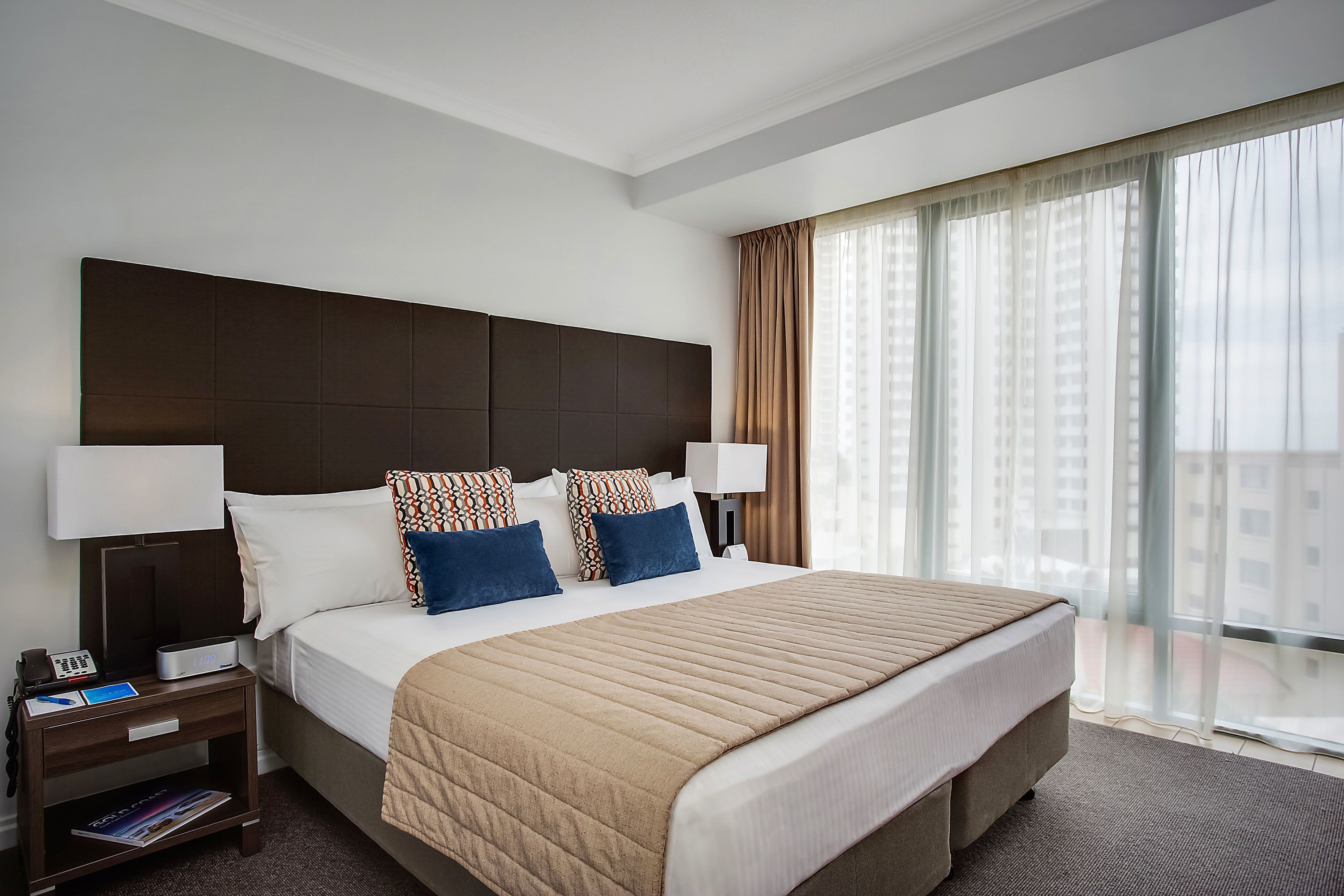Una habitación de hotel con una cama grande, una mesita de noche con un teléfono y una revista, y una ventana con cortinas.