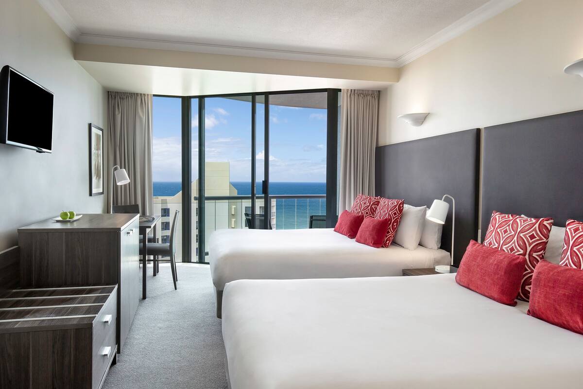 Habitación de hotel con dos camas, un escritorio, una silla, un televisor y un balcón con vista al océano.