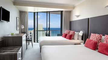 A hotel room with two beds, a desk, a chair, a TV, and a balcony with a view of the ocean.