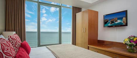 A hotel room with a large window, a bed with red and white pillows, a wooden wardrobe, a flat-screen TV, and a vase with flowers.