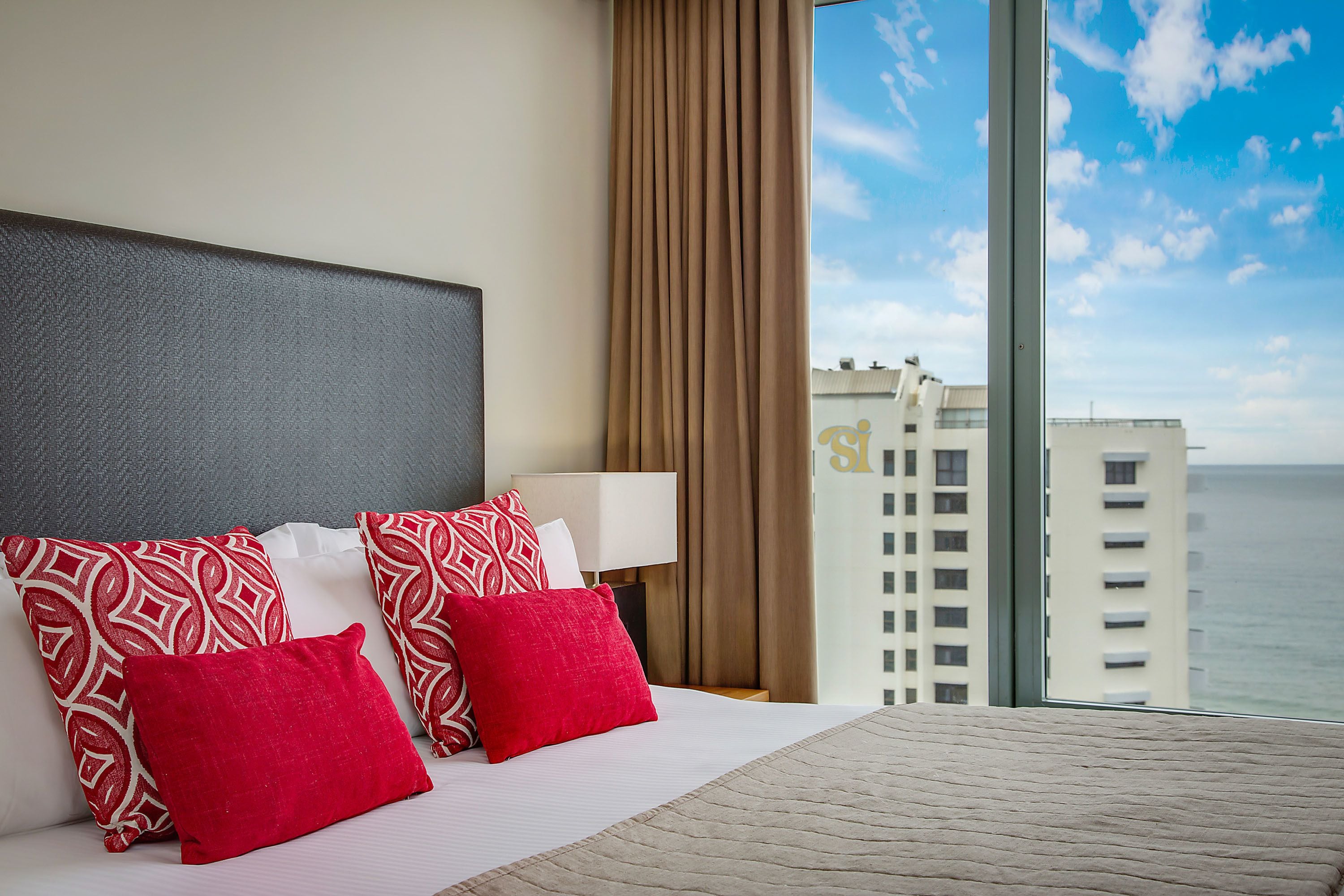 A hotel room with a large bed, red and white patterned pillows, a white headboard, a bedside lamp, and a view of the ocean and buildings.