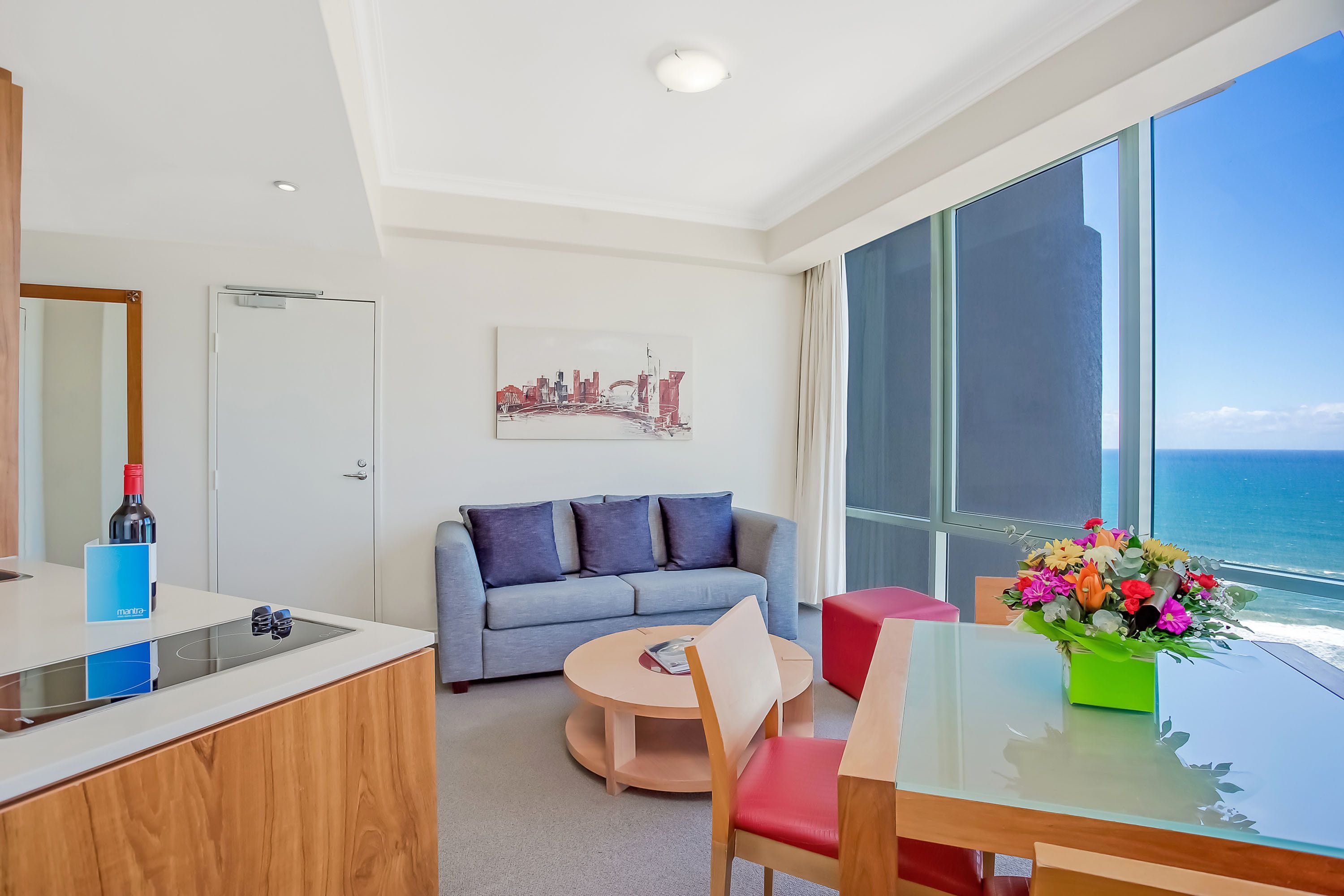 A modern hotel room with a large window, a grey sofa, a round table, and a glass dining table.