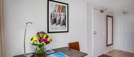 A hotel room with a wooden desk, a framed picture on the wall, a vase of flowers, and a door.