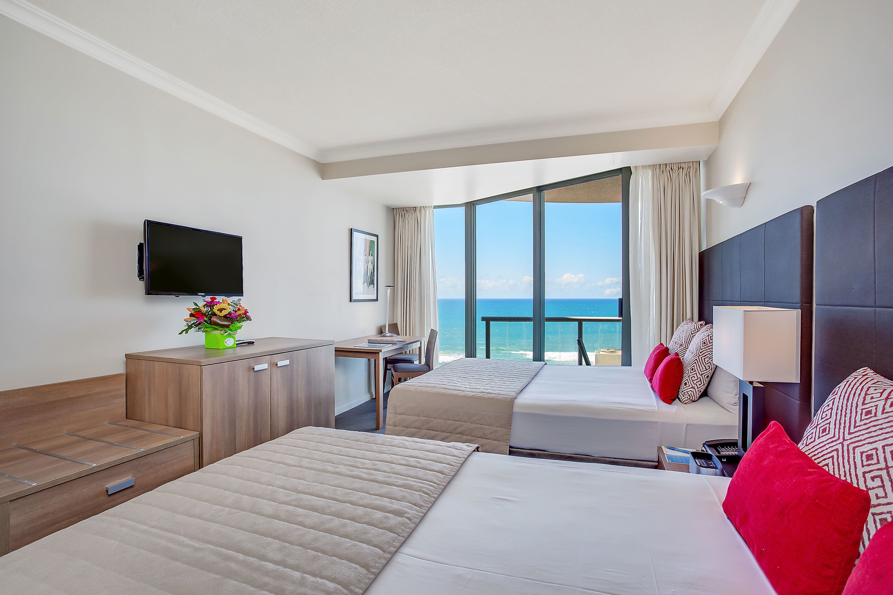A hotel room with two beds, a desk, a television, and a large window with a view of the ocean.
