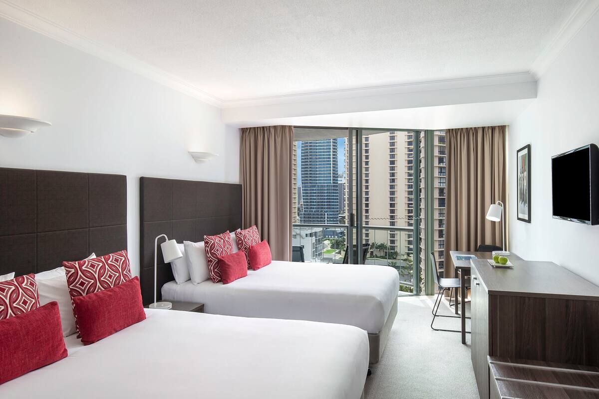 Habitación de hotel con dos camas, un escritorio, una silla y un amplio ventanal con vista a la ciudad.