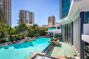 Una piscina con sillas de descanso y una cascada, rodeada de edificios altos y abundante vegetación.