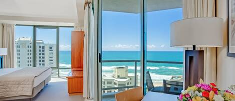 A hotel room with a bed, a desk with a lamp, a chair, and a view of the ocean through large glass doors.