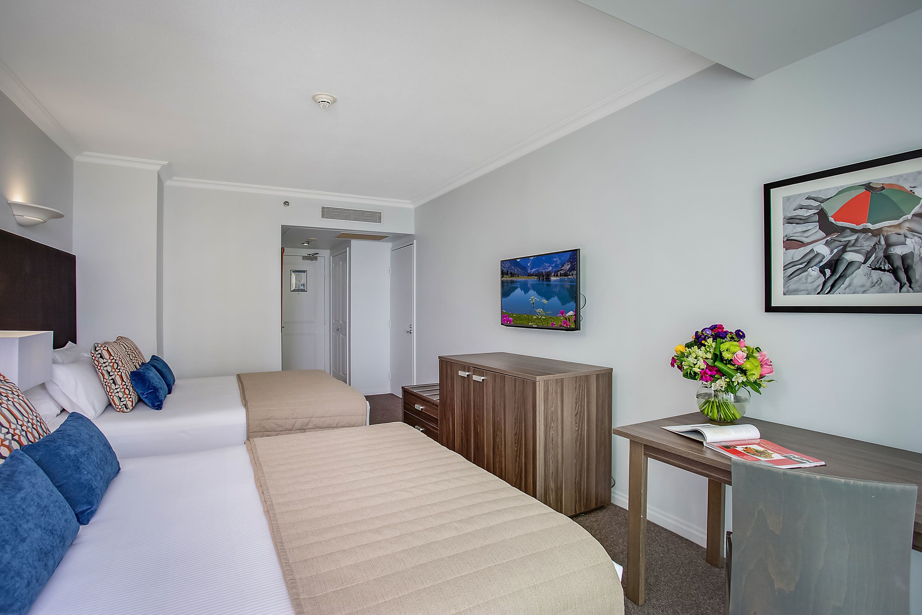 A hotel room with two beds, a wooden cabinet, a desk with a vase of flowers, and a framed picture on the wall.