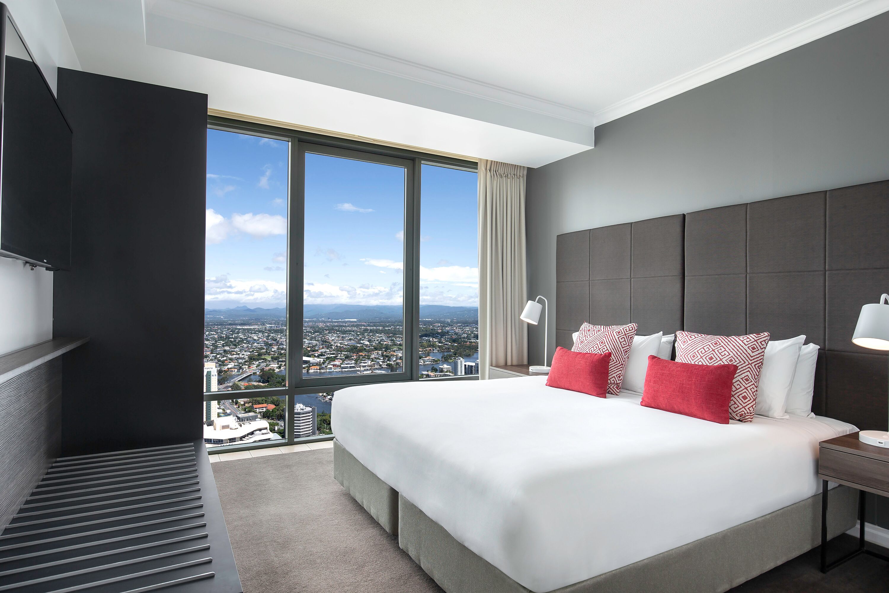 A modern hotel room with a large bed, a bedside table, a nightstand, and a view of the city through large windows.