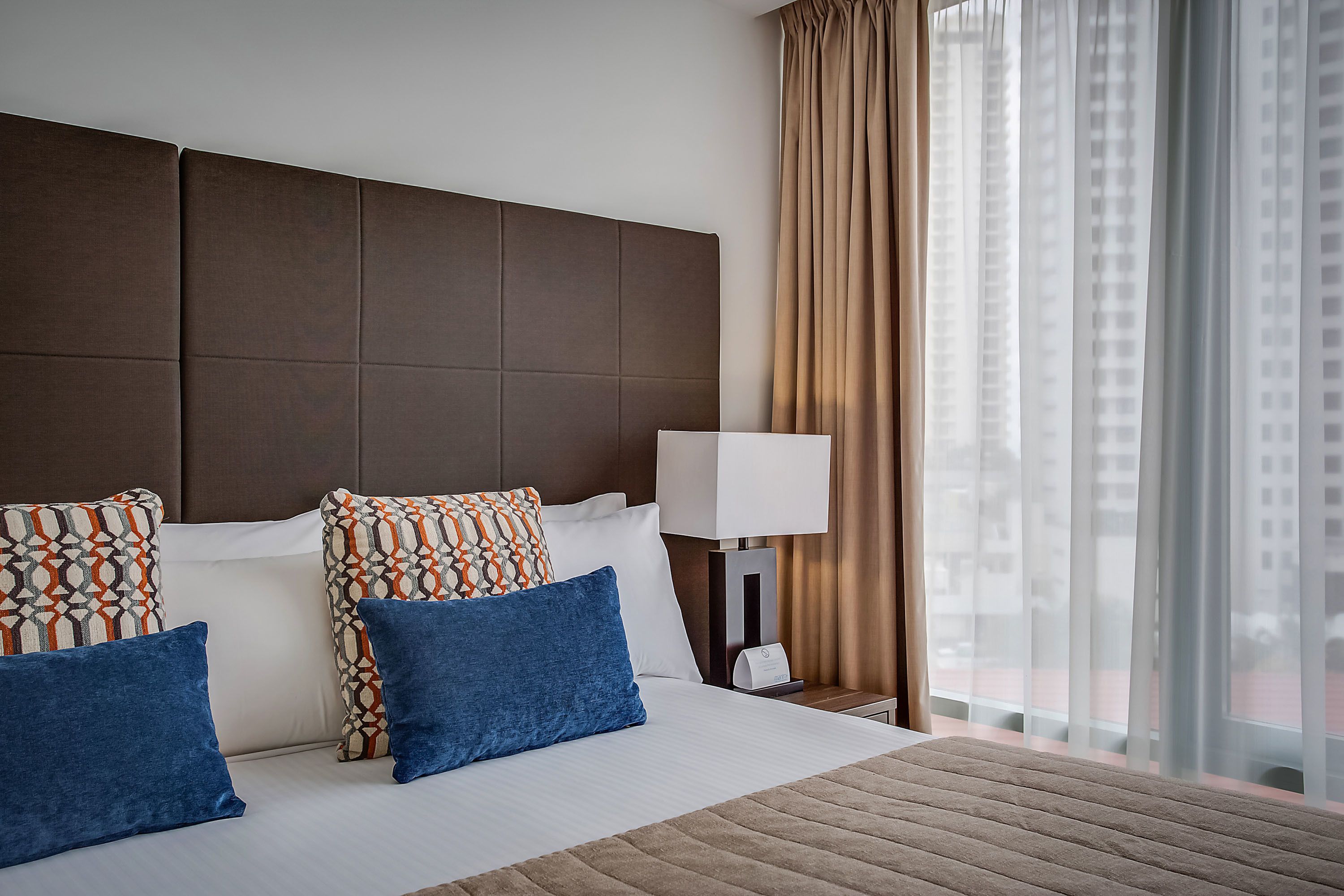 Une chambre d’hôtel avec un grand lit, de nombreux oreillers, une table de chevet avec une lampe et une vue sur la ville à travers de fines rideaux.