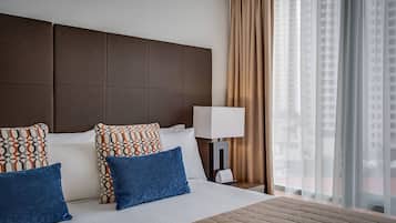 Habitación de hotel con una cama grande, varios almohadones, una mesita de noche con lámpara y vistas a la ciudad a través de cortinas traslúcidas.