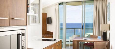 A modern hotel room with a kitchen, a dining area with a table and chairs, a TV, and a balcony with a view of the ocean.