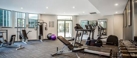 A modern gym with various exercise equipment, large windows, and a carpeted floor.