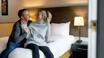 A couple sitting on a bed in a hotel room with a nightstand and a lamp.