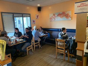 A restaurant interior with wooden tables and chairs, a blue cushioned bench, and a painting of flowers on the wall.