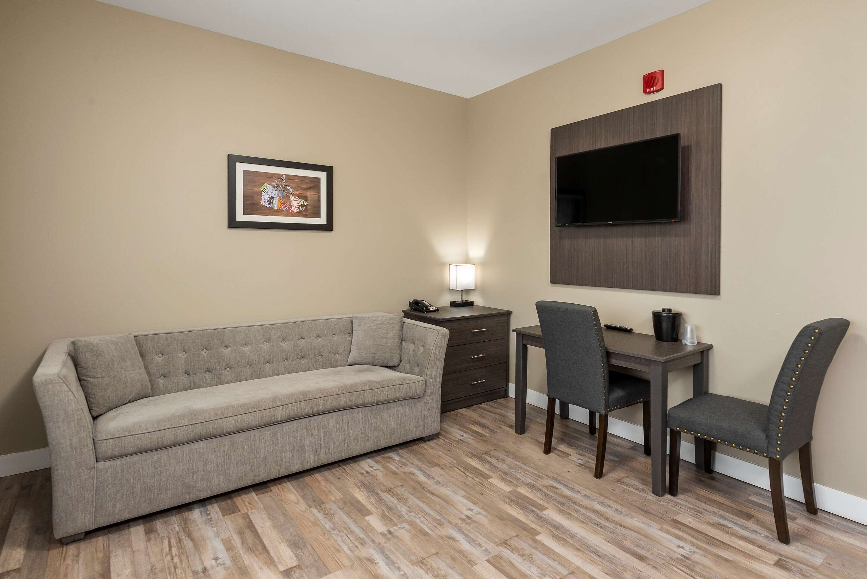 Un salon comprenant un canapé, un bureau, deux fauteuils, un téléviseur à écran plat fixé sur un panneau en bois, un tableau encadré au mur et une petite table avec une lampe.