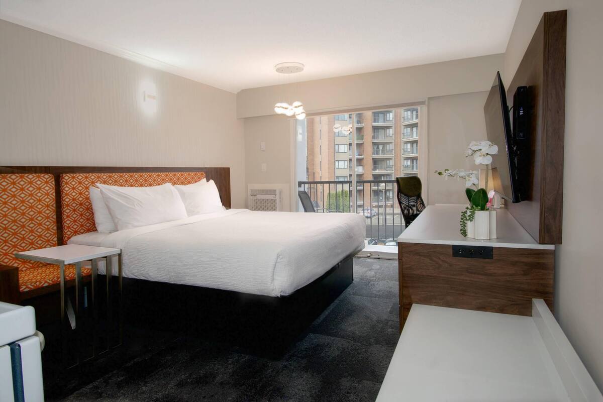 A hotel room with a bed, a small table, a desk, a mirror, and a balcony with a view of buildings.