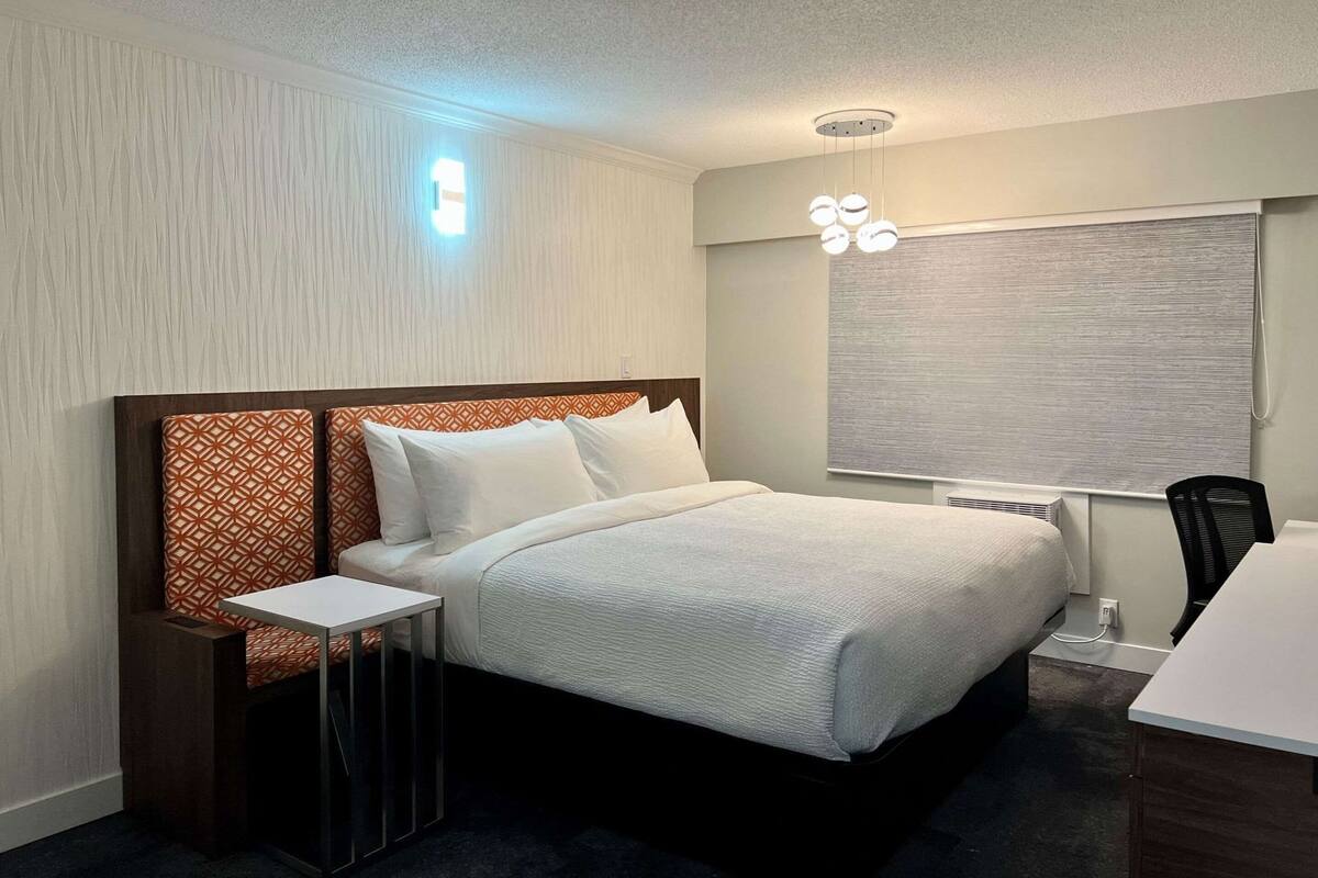A hotel room with a bed, bedside table, chair, and a window with blinds.