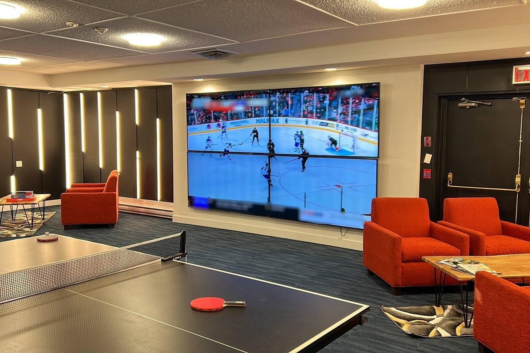 Camera d'albergo con una grande TV che mostra una partita di hockey, poltrone rosse, un tavolo da ping pong e una porta con il segnale di uscita.