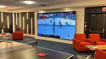 Camera d'albergo con una grande TV che mostra una partita di hockey, poltrone rosse, un tavolo da ping pong e una porta con il segnale di uscita.