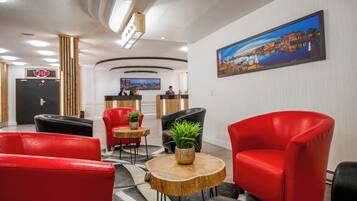 A hotel lobby with red leather chairs, a reception desk, a large cityscape painting, and a wooden coffee table with a plant.
