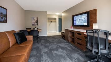 Una sala moderna con un sofá de cuero marrón, una mesa de comedor con sillas, un escritorio de madera con computadora y silla, y un televisor montado en la pared.