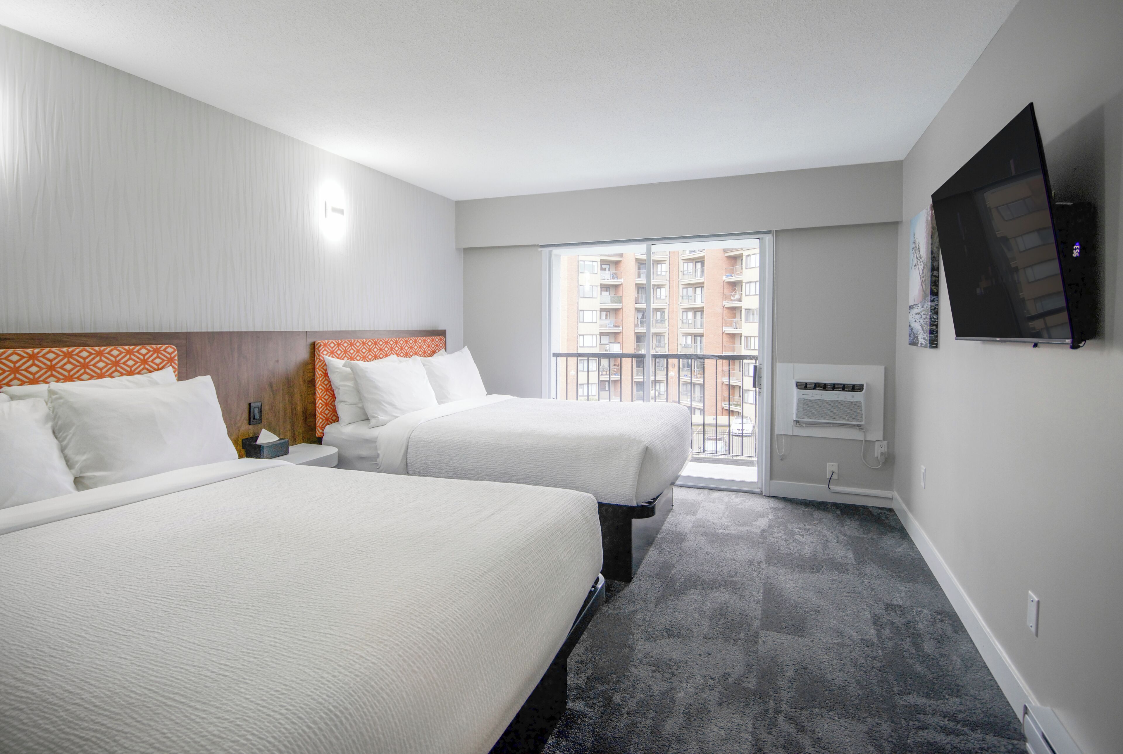 A hotel room with two beds, a TV, a balcony with a view of buildings, and a carpeted floor.