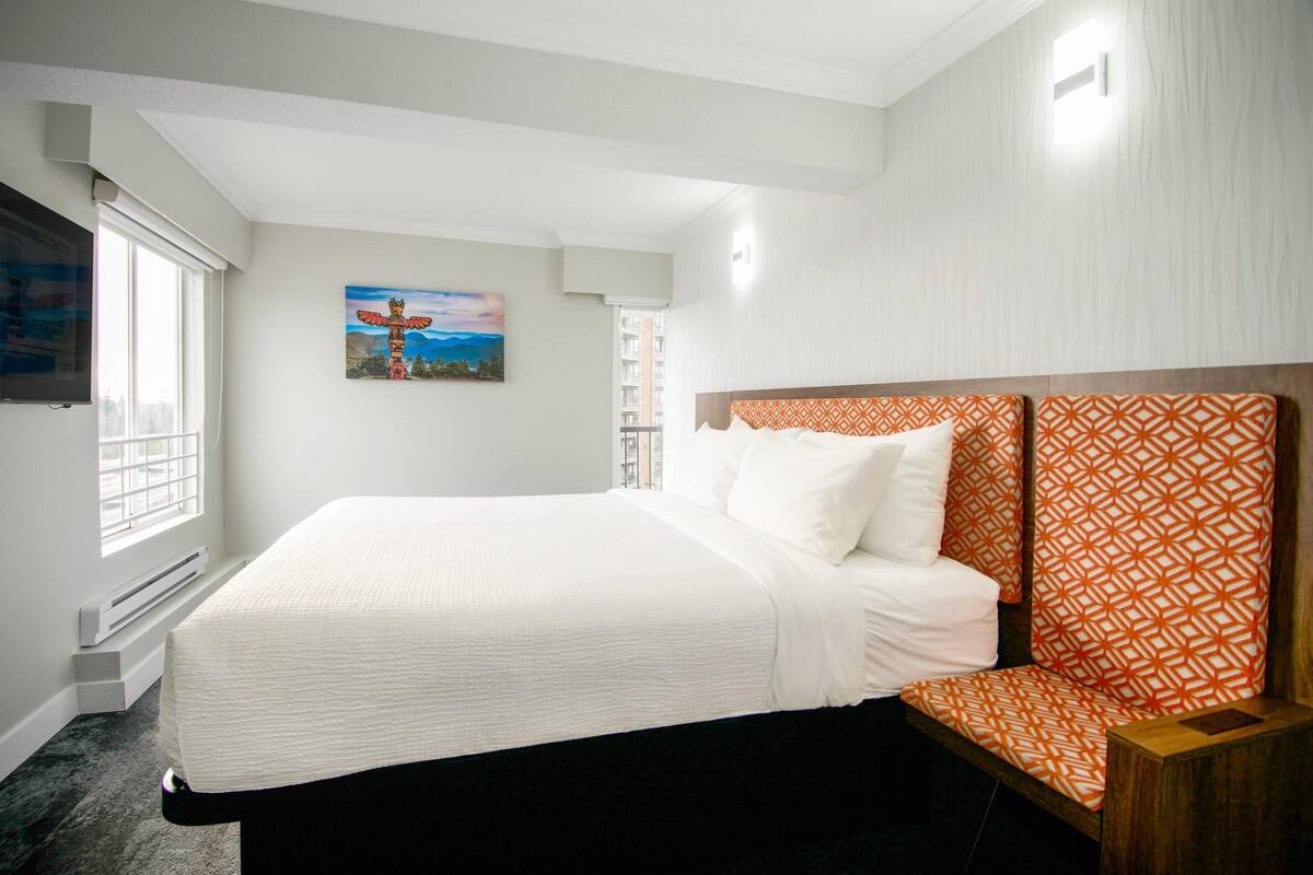 A hotel room with a large bed, a headboard with a geometric pattern, a painting on the wall, and a window with a view of buildings.