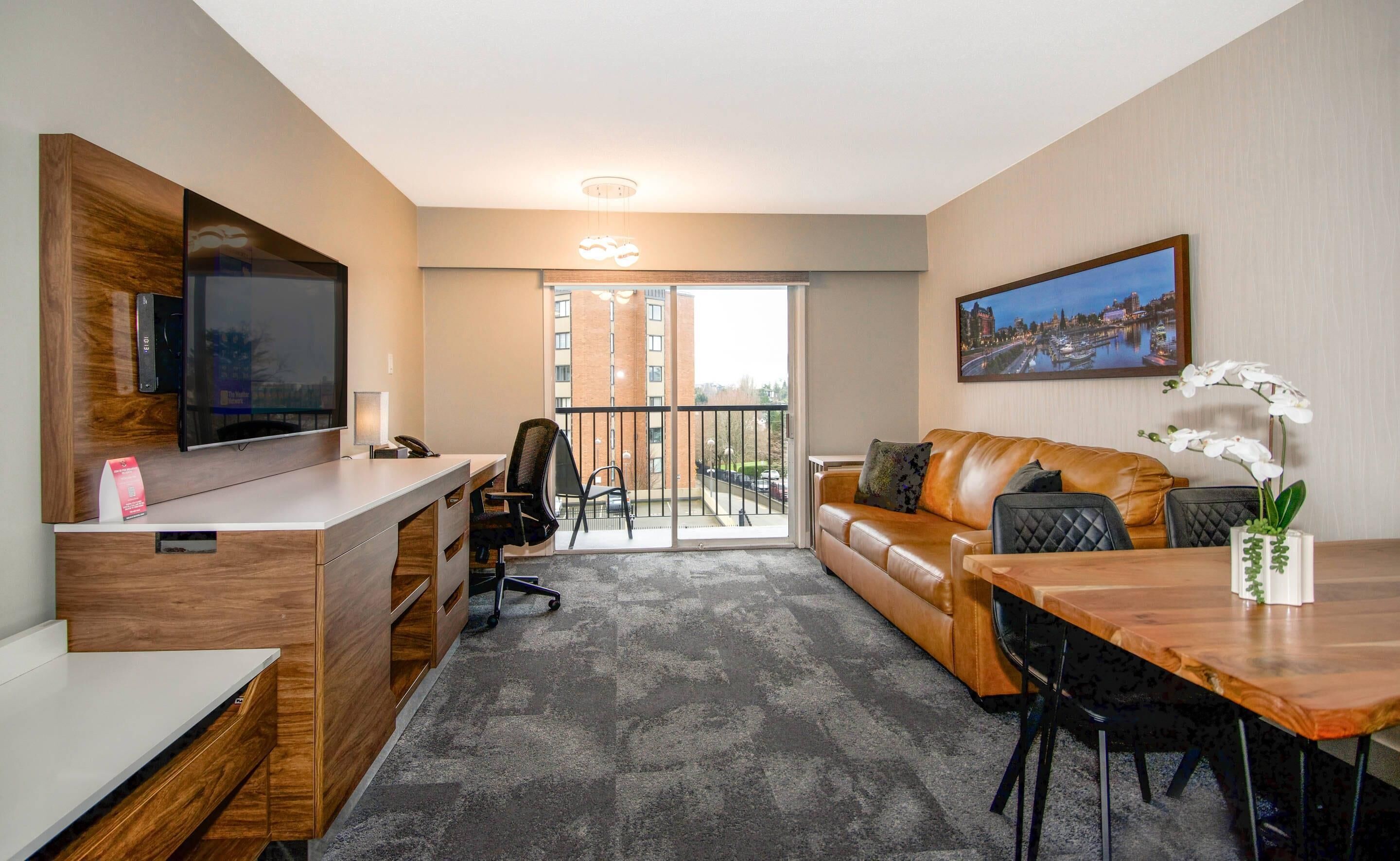 Quarto de hotel moderno com televisão de tela plana, escrivaninha com telefone, sofá de couro, mesa de jantar com cadeiras e varanda com vista para a cidade.
