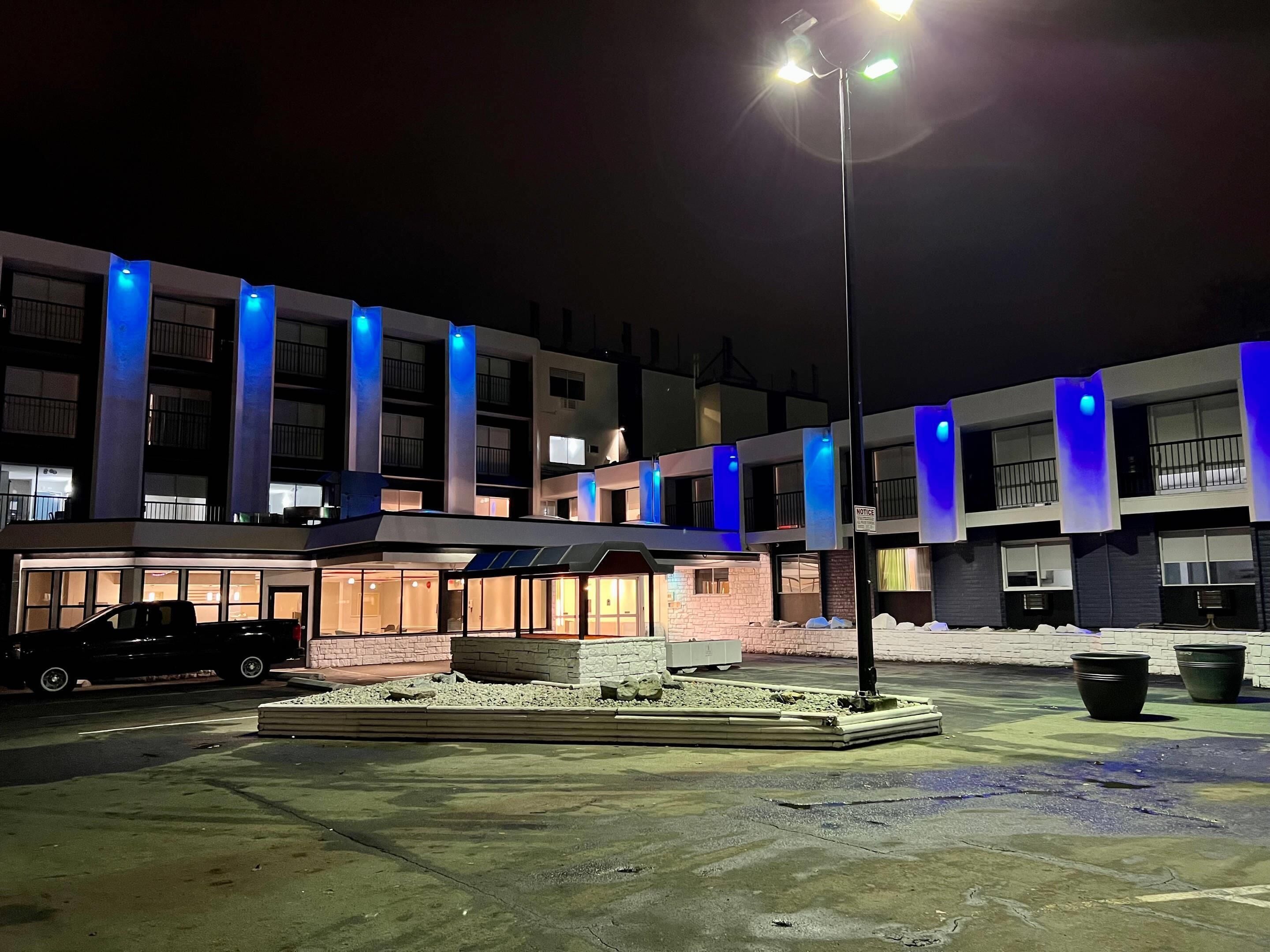 A multi-story hotel with illuminated vertical columns, a parking area, and a small outdoor seating area.