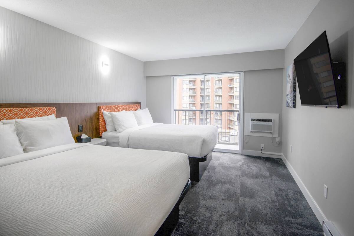A hotel room with two beds, a TV, a balcony, and a view of a building.
