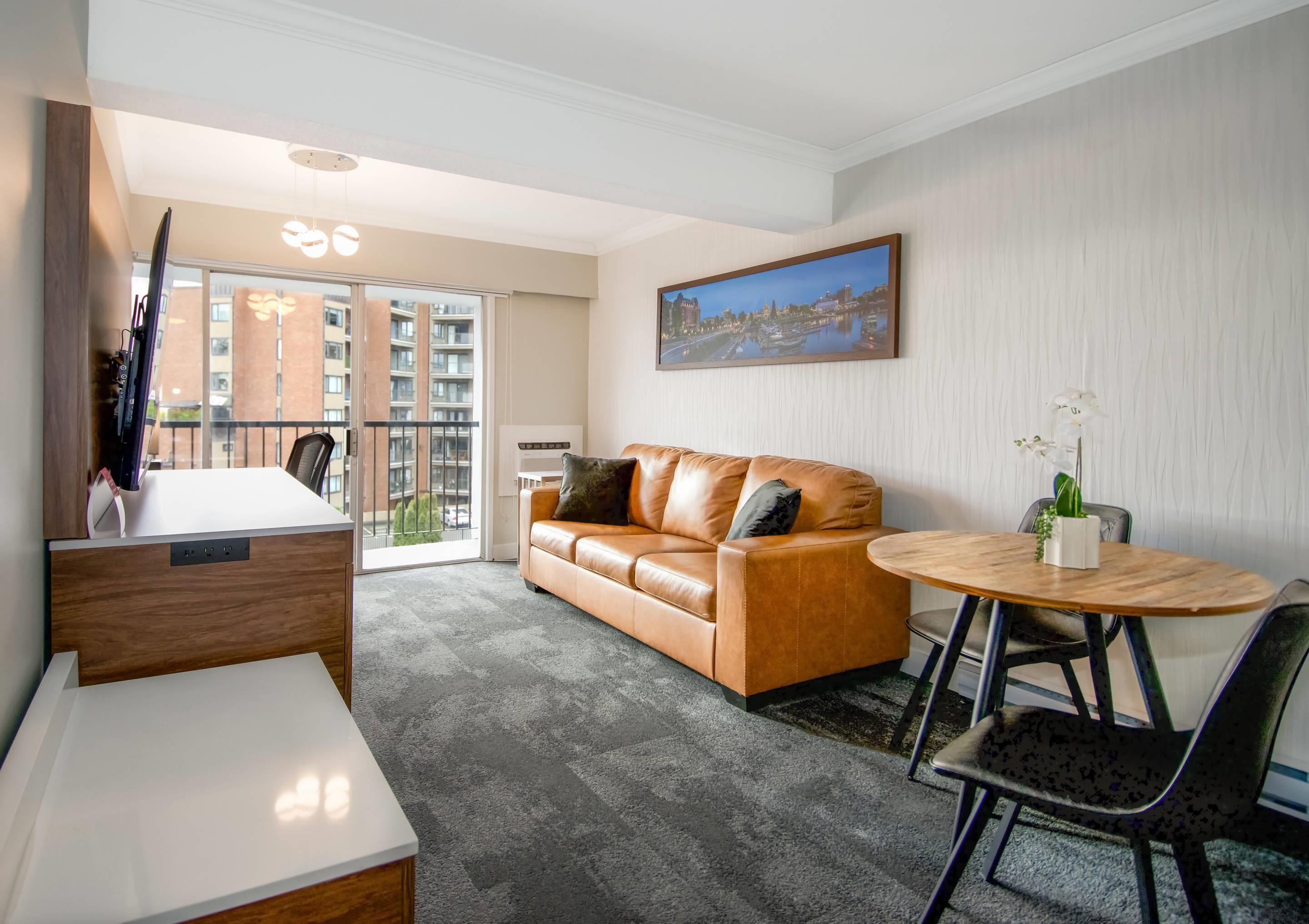 A modern hotel room with a leather sofa, a round wooden table, and a large window offering a city view.