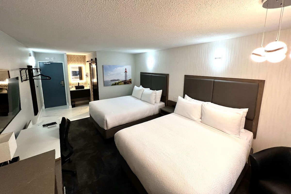A hotel room with two beds, a desk, a chair, a mirror, a door, and a painting of a lighthouse.