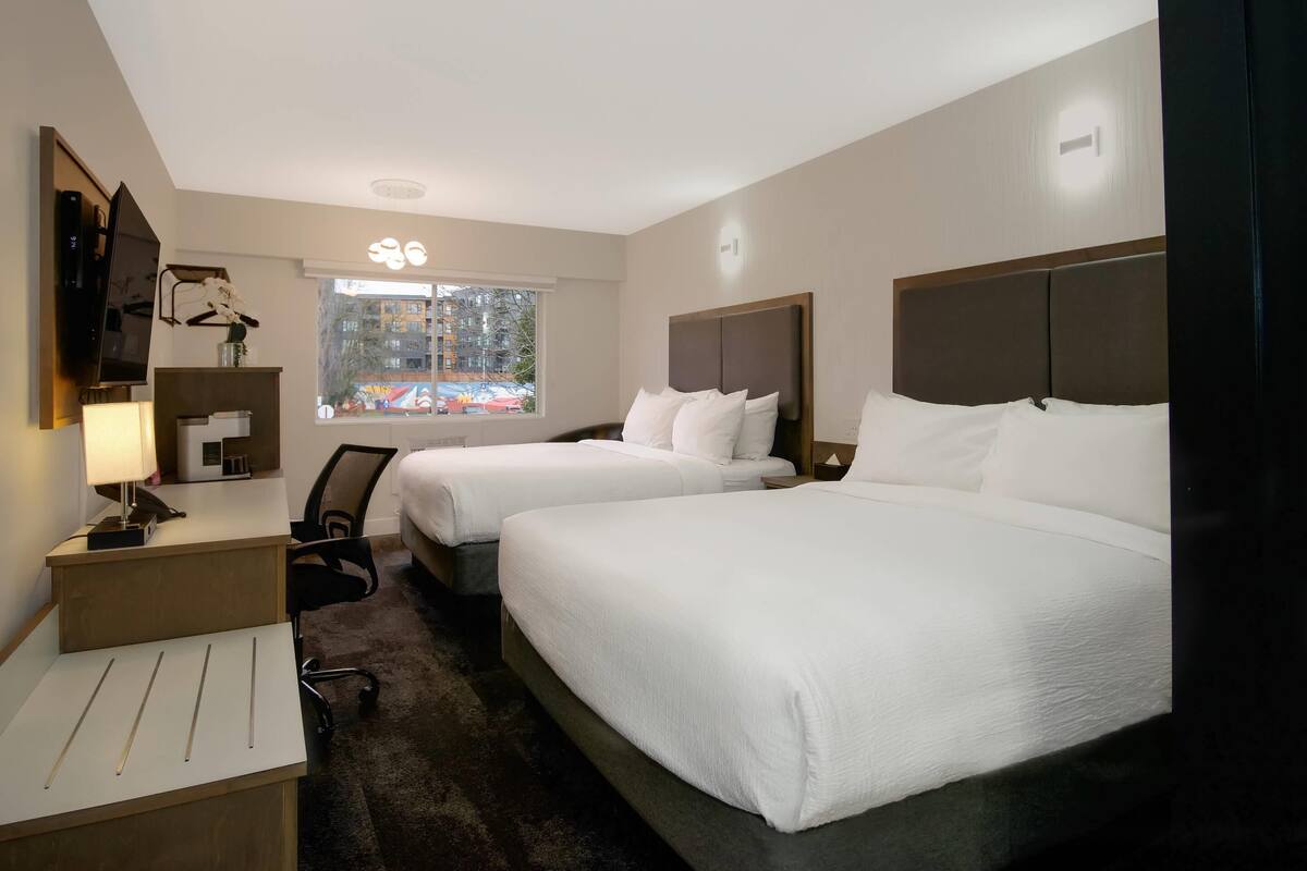 A hotel room with two beds, a desk, a chair, a TV, and a window with a view of buildings.