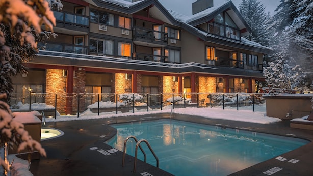A multi-story hotel with a pool area, surrounded by snow-covered trees and a brick facade. - Whistler Village Inn And Suites (Whistler)