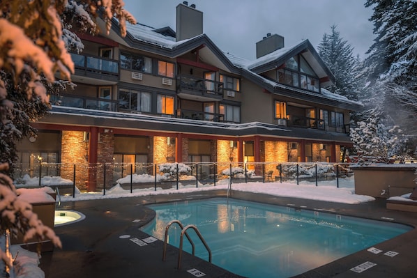 A multi-story hotel with a pool area, surrounded by snow-covered trees and a brick facade. - Whistler Village Inn And Suites (Whistler)