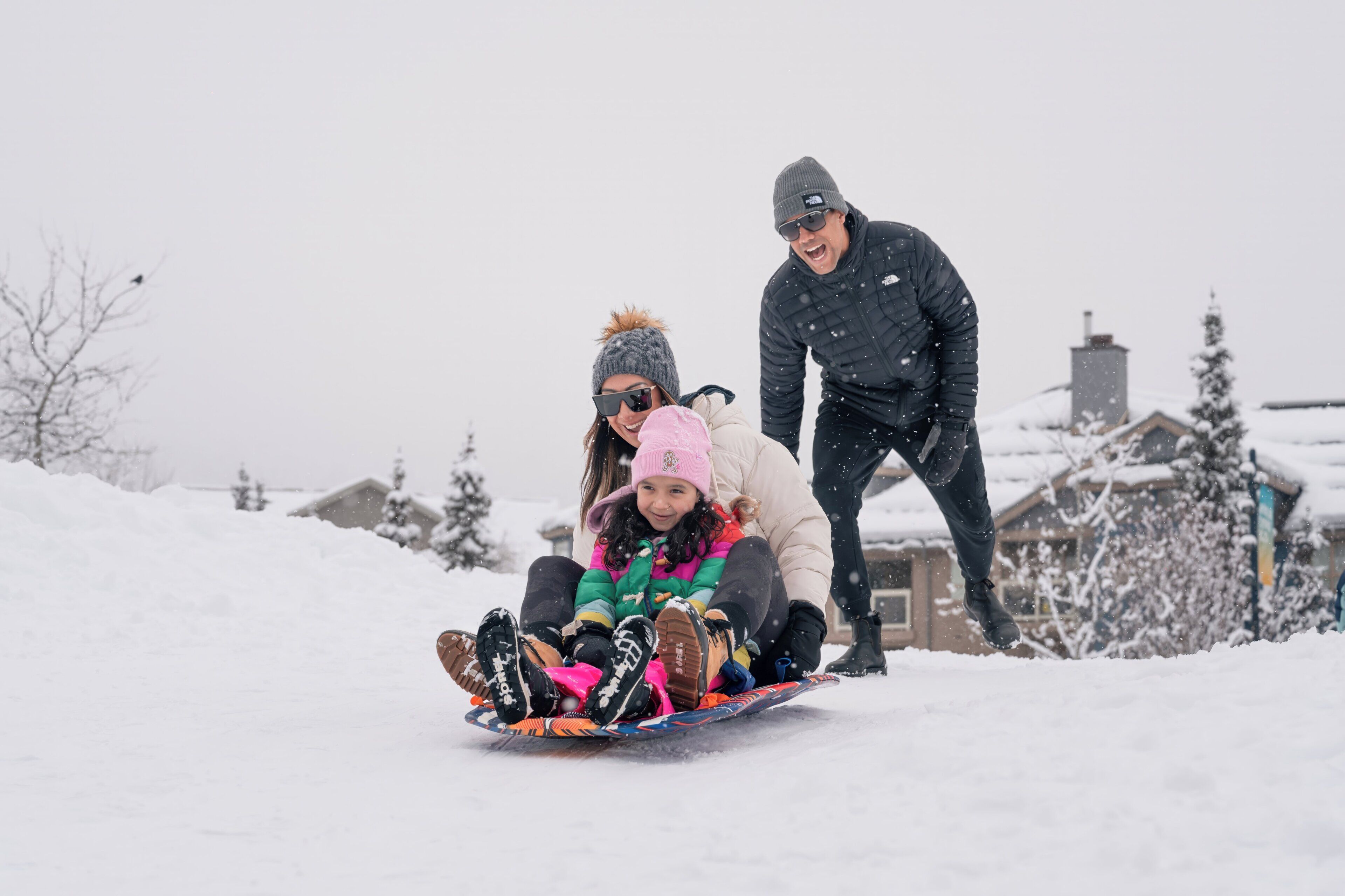 A snowy outdoor scene with a person sledding, another person standing, and a child in a colorful outfit.