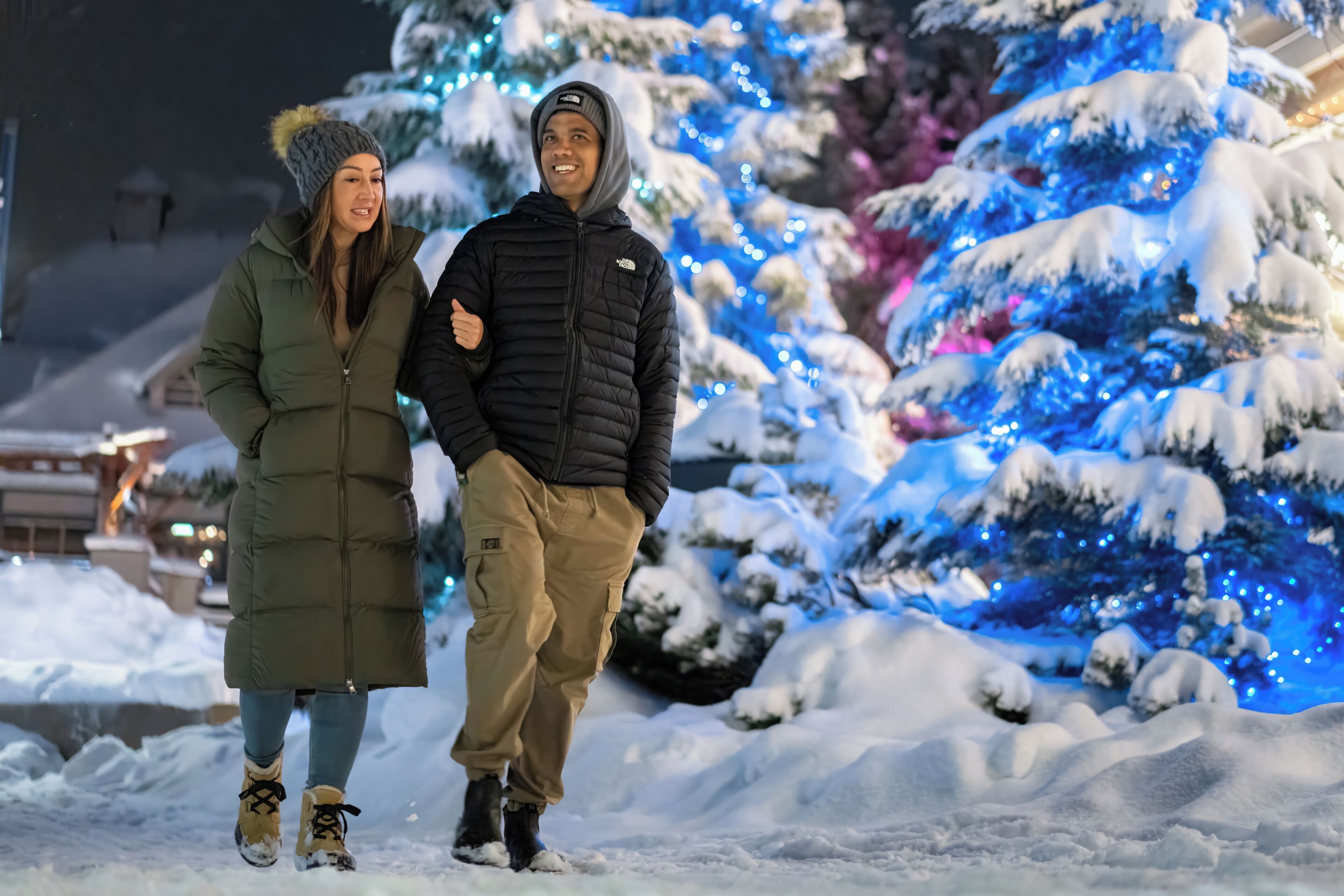 Twee personen lopen door een besneeuwde omgeving met sneeuw bedekte bomen en blauw licht.