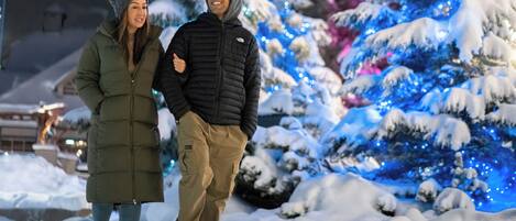 Two people walking in a snowy outdoor area with snow-covered trees and blue lights.
