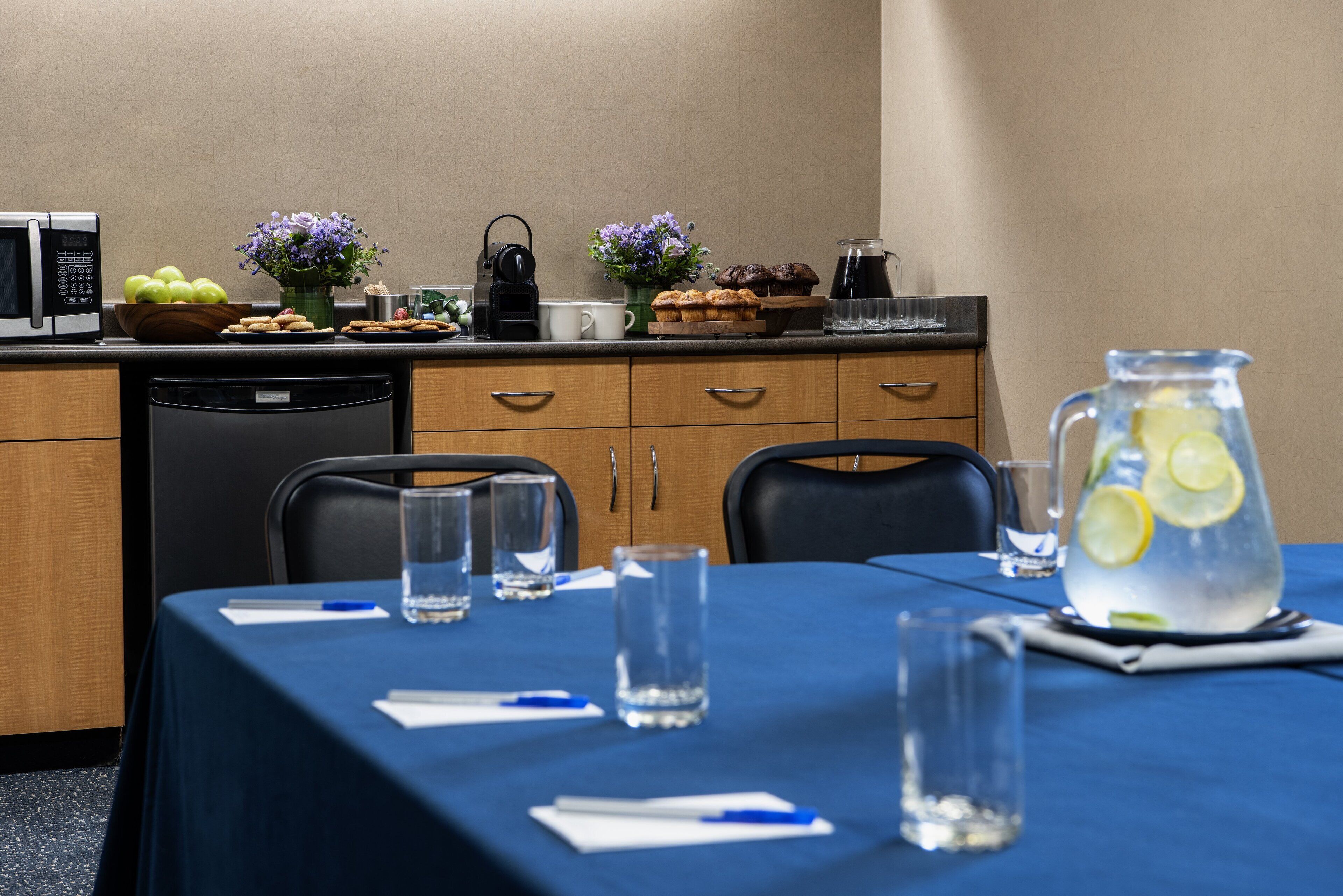 Un salón de conferencias con mantel azul, vasos de agua, jarrón de limonada y microondas sobre la encimera.