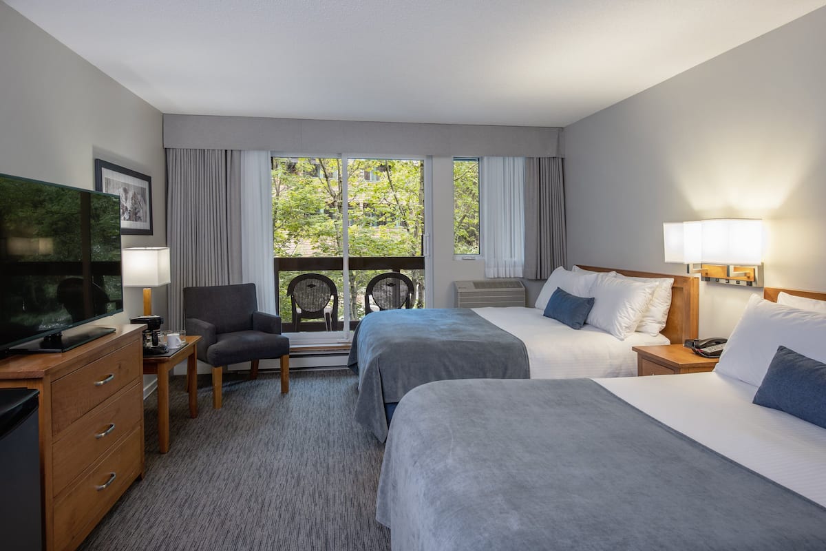 A hotel room with two beds, a TV, a desk with a lamp, a chair, and a view of trees through large windows.