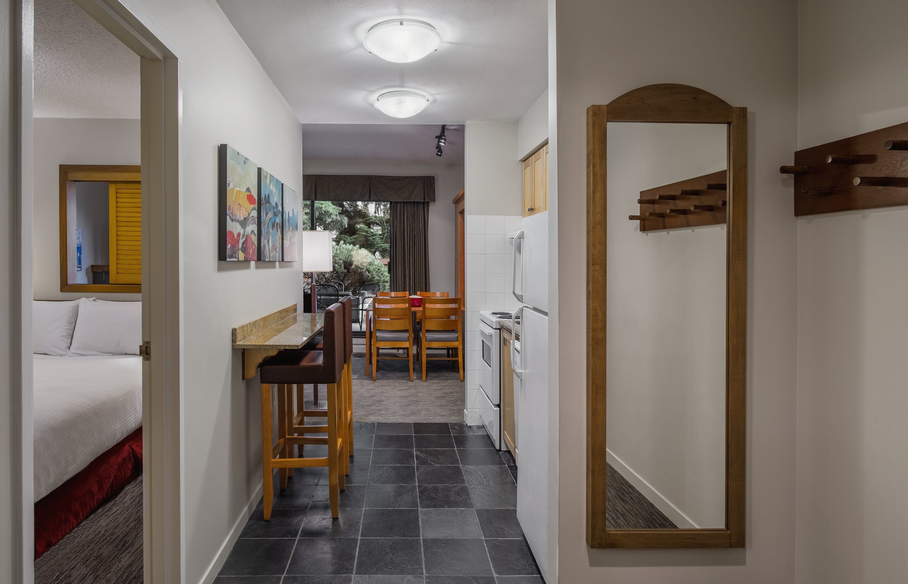 A narrow hallway leading to a bedroom with a bed, a small dining area with a table and chairs, a kitchen with a refrigerator and stove, and a large mirror on the right wall.