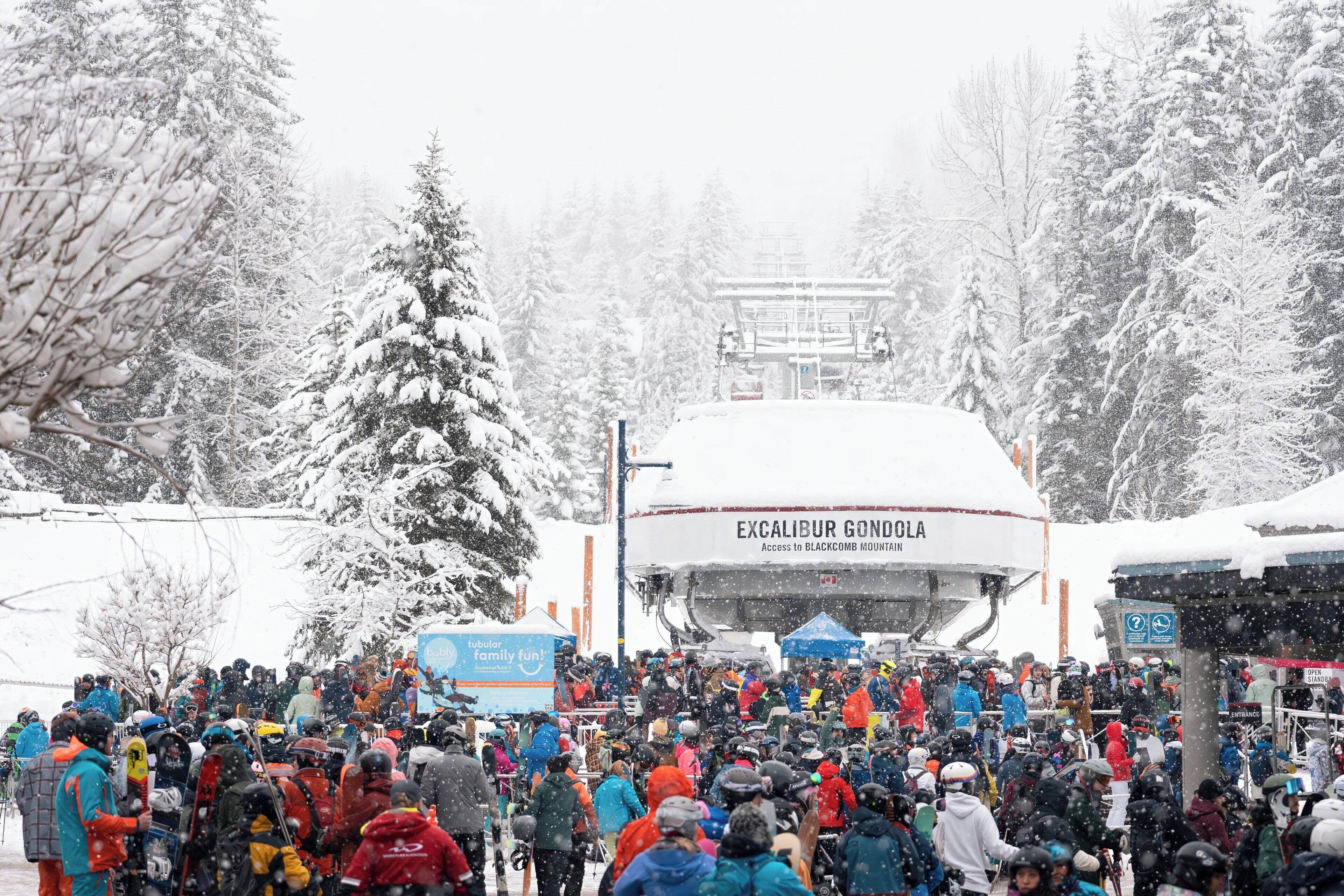 A snowy outdoor scene with a crowd of people, some with snowboards and skis, and a large sign for Excaliibur Gondola.