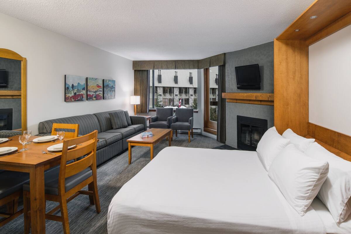 A hotel room with a bed, a sofa, a dining table with chairs, a fireplace, a television, and artwork on the wall.