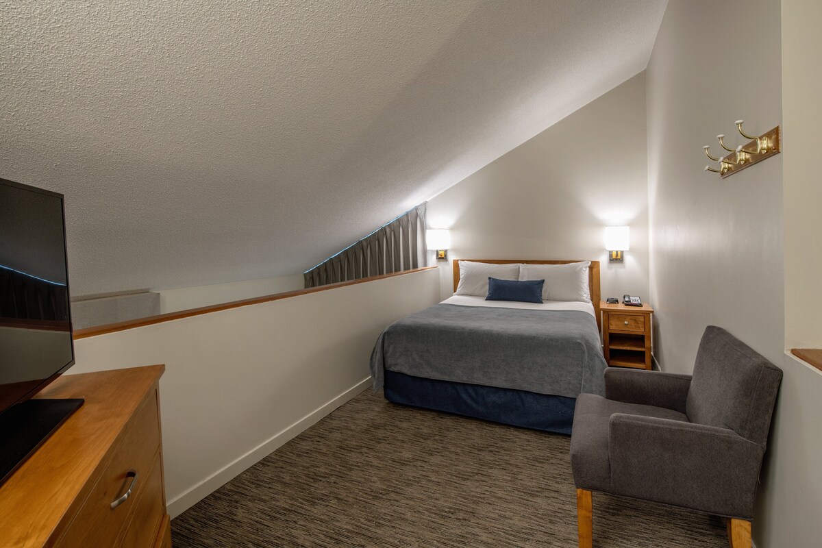 A bedroom with a bed, a chair, a TV, a nightstand, and a wall-mounted light fixture.