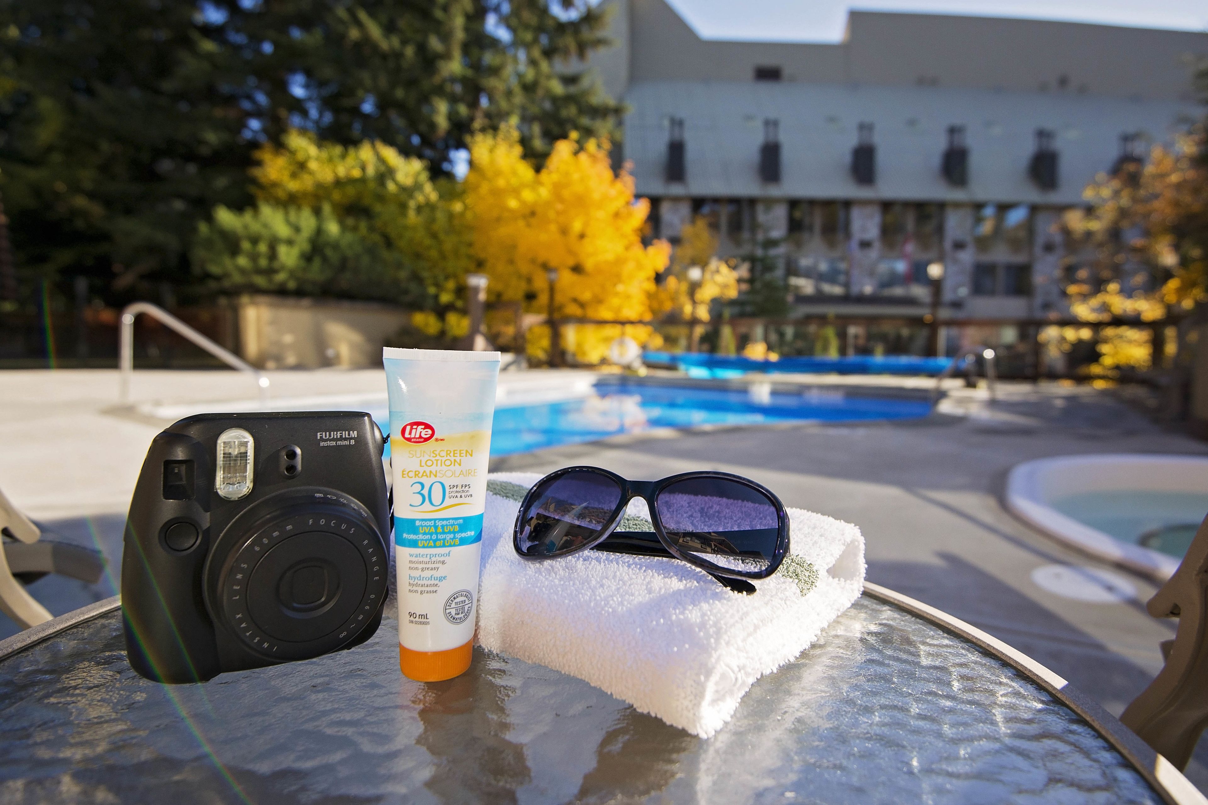 Un appareil photo, de la crème solaire et des lunettes de soleil sont posés sur une table près d’une piscine, avec un bâtiment en arrière-plan.