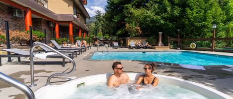 A hot tub area with a pool and lounge chairs, surrounded by trees and a building with balconies.