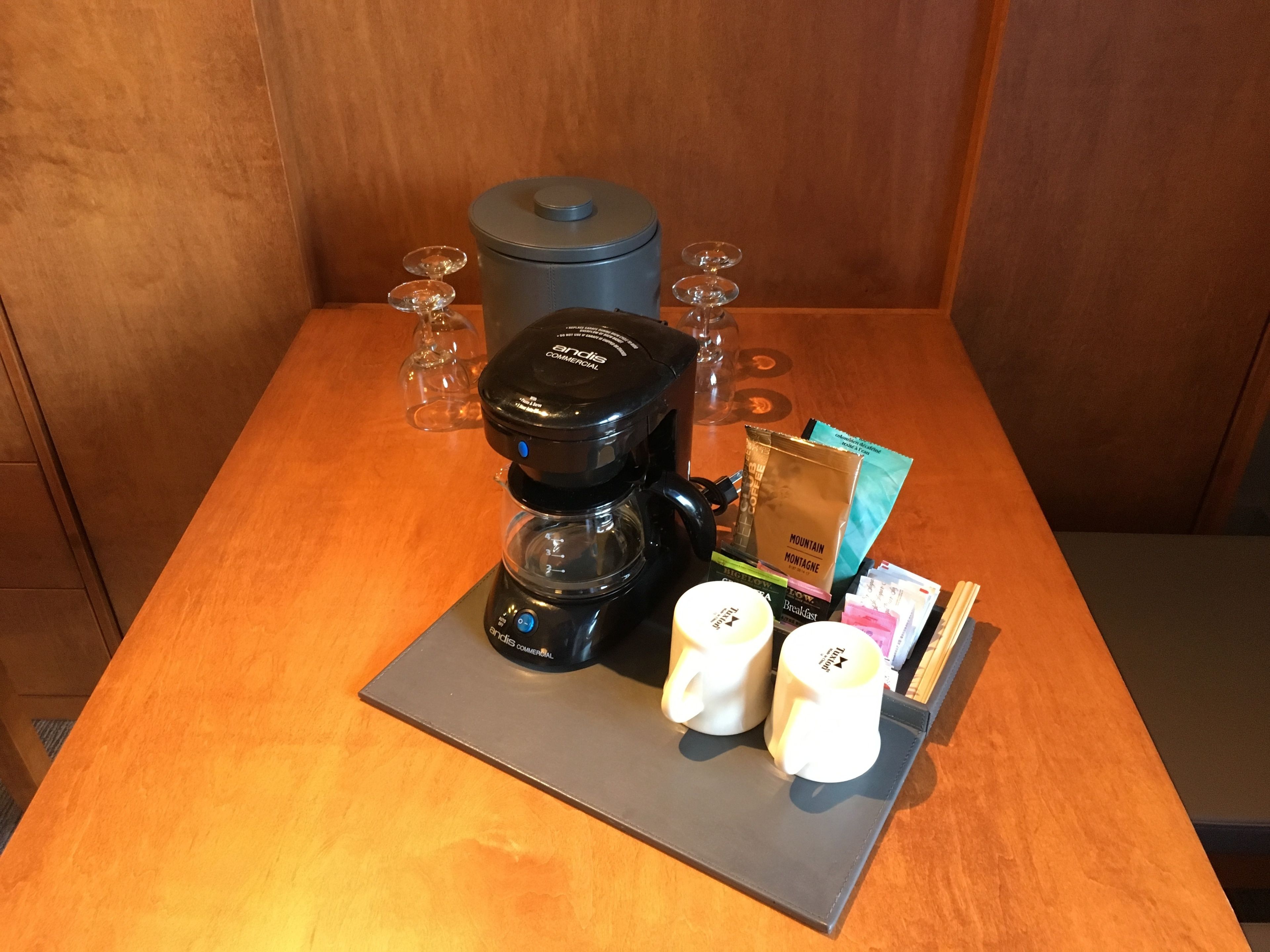 Eine Kaffeemaschine, zwei weiße Tassen, eine Kaffeepackung und ein Glas Wasser auf einem Holztisch.