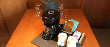 Una cafetera, dos tazas blancas, un paquete de café y un vaso de agua sobre una mesa de madera.