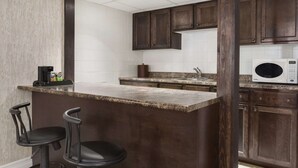 A kitchen with dark wooden cabinets, a granite countertop, a white microwave, and two black bar stools.